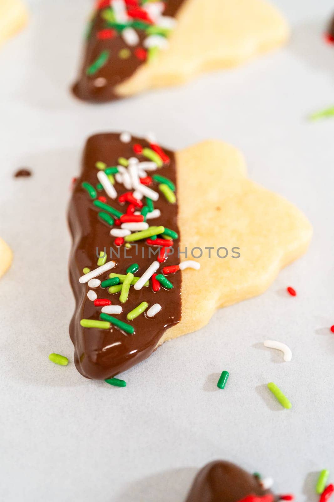 Buttery cookies half-dipped in chocolate, sprinkled with festive red, green, and white decorations, laid on white parchment to set.