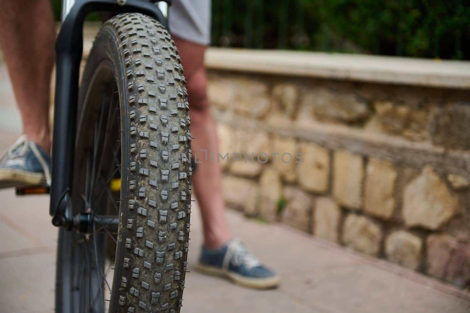 Shallow tread of a bicycle tubeless tire. Selective focus on e-bike wheel and feet on pedals of a male cyclist in denim sneakers by artgf