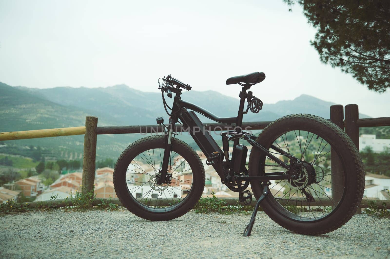 A modern black electric bike on the countryside road, parked by a wooden fence, against mountains background in the nature. Copy advertising space. Environmental conservation. Sustainable lifestyle