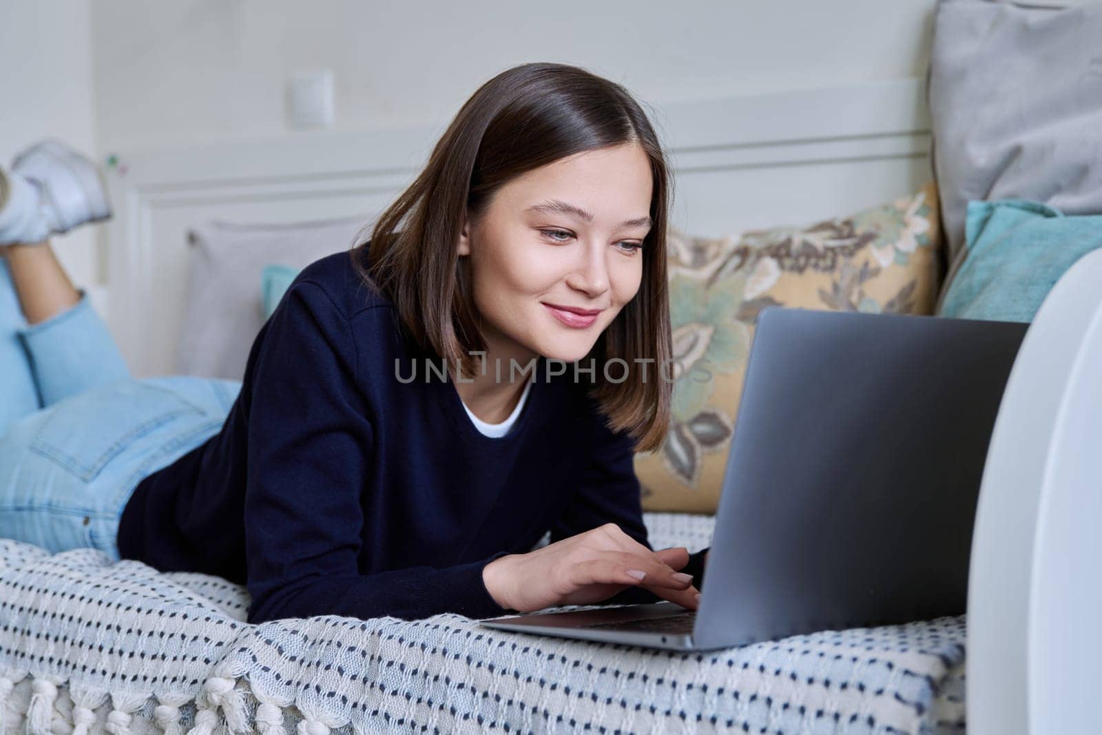 Young woman using laptop computer, typing on keyboard, lying on sofa at home. Internet online technologies for work communication study leisure