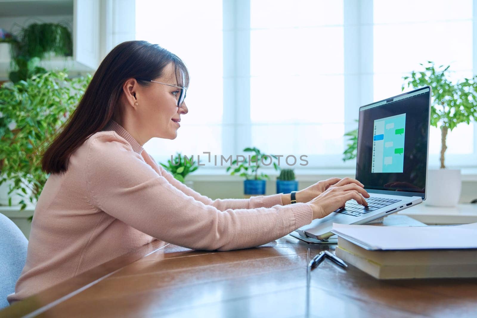 Middle-aged woman communicates online using laptop at desk at home by VH-studio