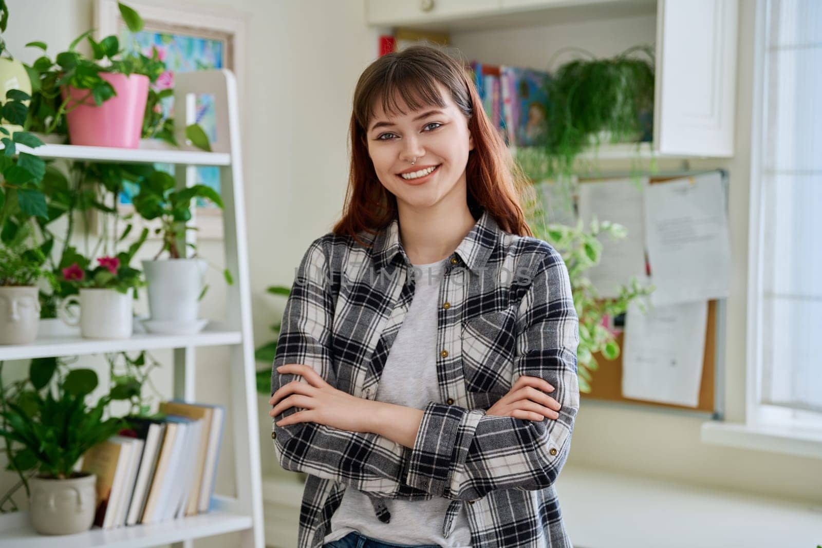 Portrait of female teenager 18-20 years old, smiling confident looking at camera with crossed arms in home. Beautiful girl with nose piercing, trendy makeup hairstyle. Youth lifestyle beauty fashion