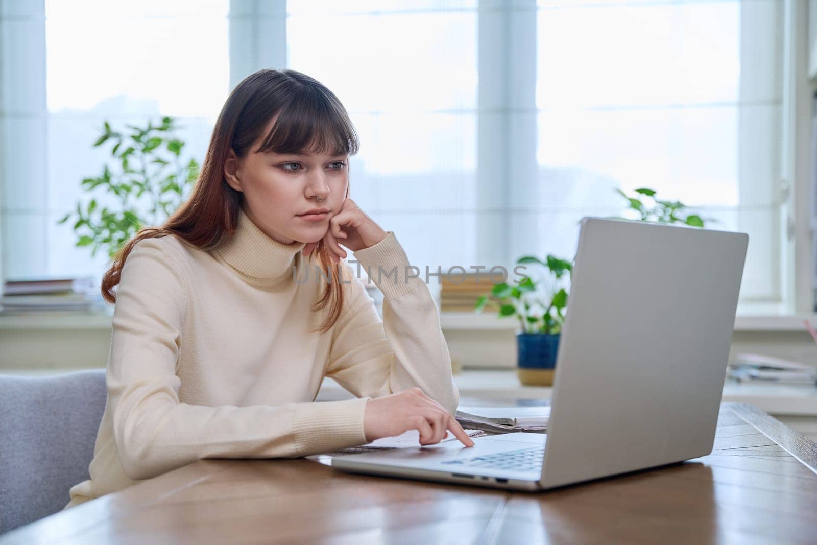 College student girl sitting at desk using laptop computer, typing on a keyboard, at home. Teenager female watching webinar, preparing for exam tests, studying remotely.