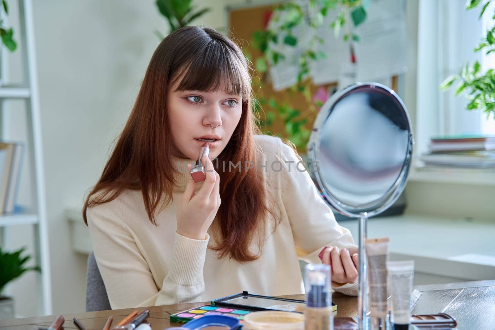 Young woman with cosmetic products on table, doing makeup looking in mirror at home, painting lips with lipstick. Cosmetics, fashion, youth, lifestyle, beauty concept