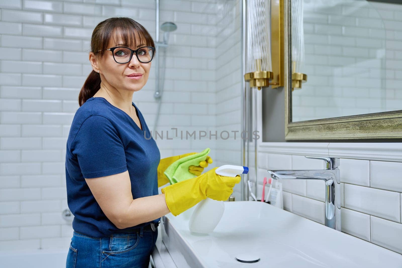 Smiling woman with detergent spray washcloth cleaning bathroom by VH-studio