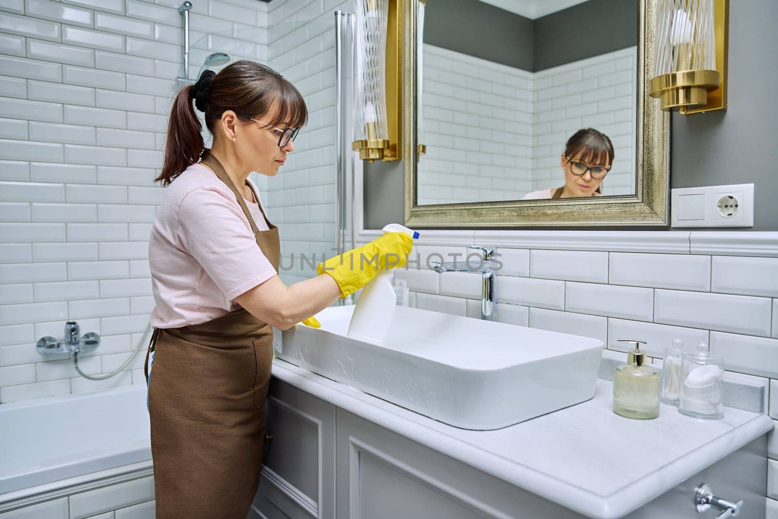 Woman in apron with detergent washcloth cleaning in bathroom, washing sink washbasin. Housewife cleaning house, cleaner service worker at workplace. Home hygiene housecleaning housekeeping housework