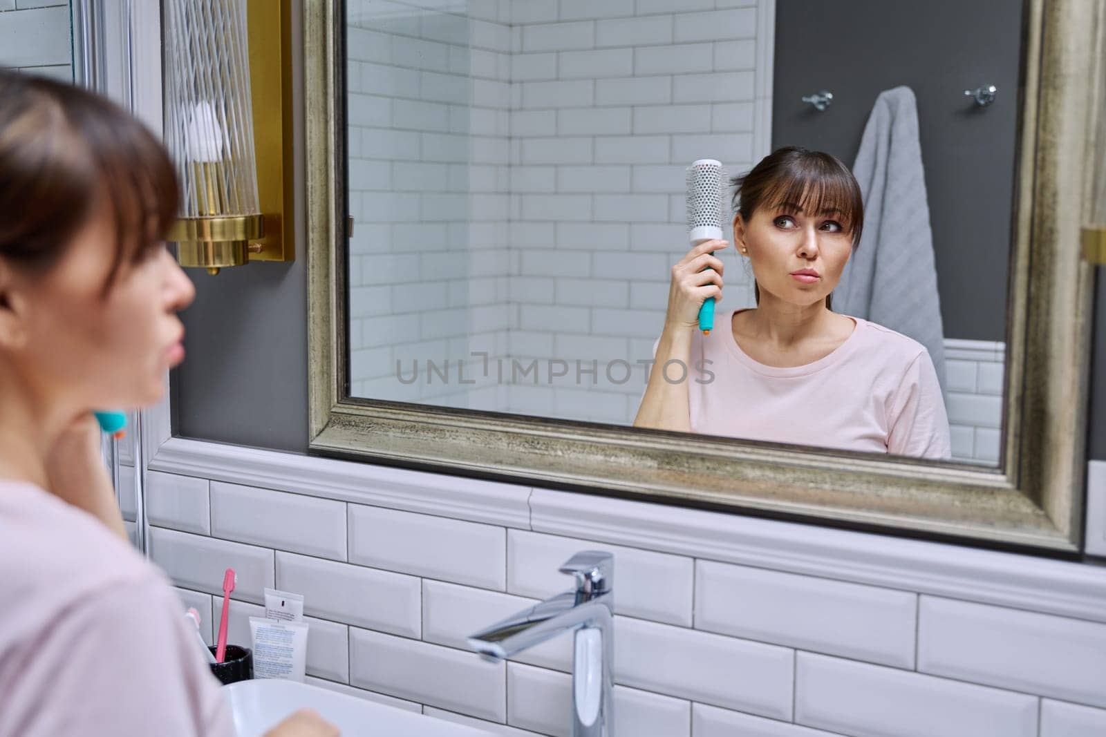Middle-aged woman with comb looking in mirror, doing hair styling, in bathroom. Beauty, age, cosmetics, hair accessories