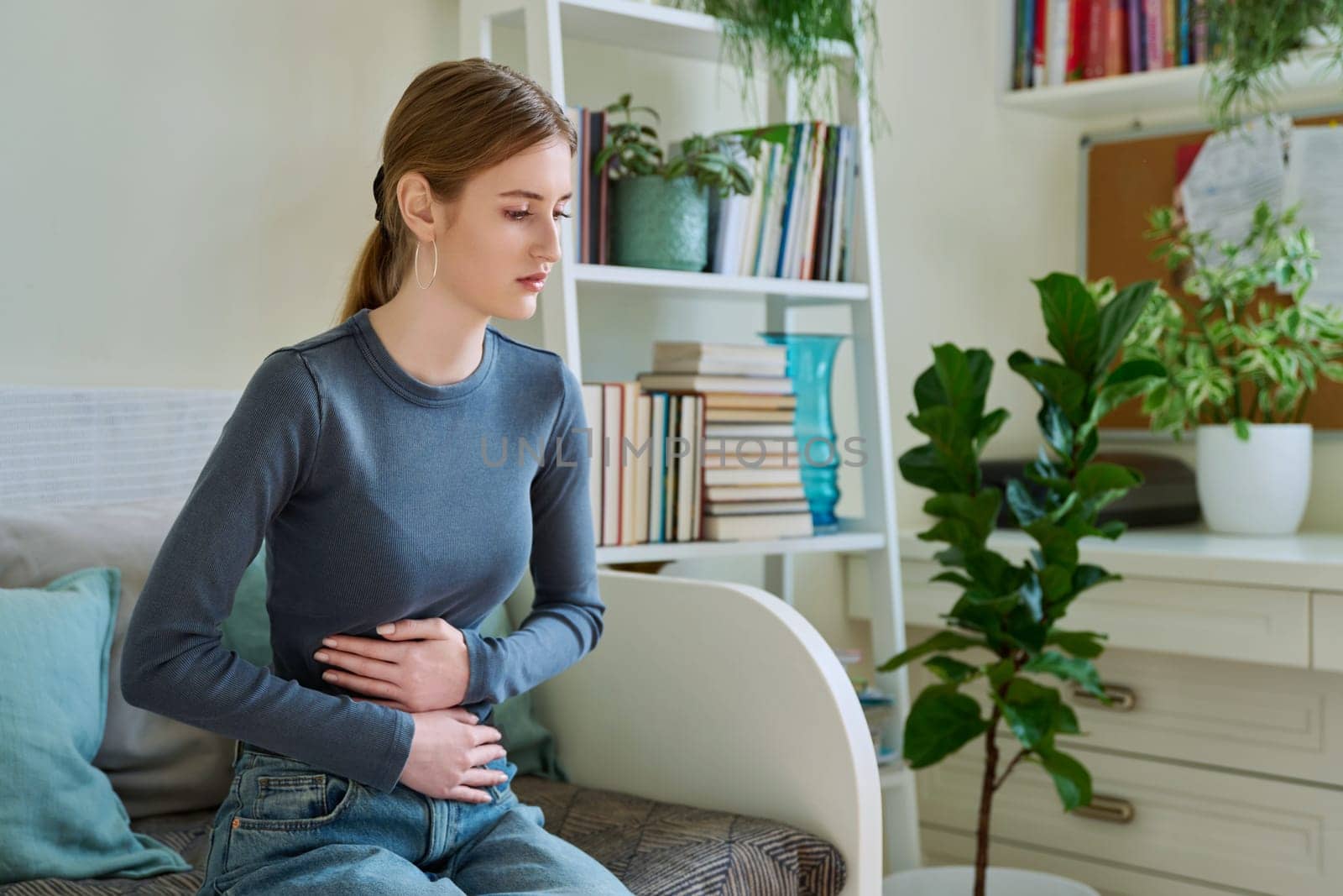 Young teenage female experiencing abdominal pain sitting at home. Stomach, menstrual cramps, gastrointestinal tract diseases, inflammatory processes in abdominal organs, kidneys, pelvis, youth, health