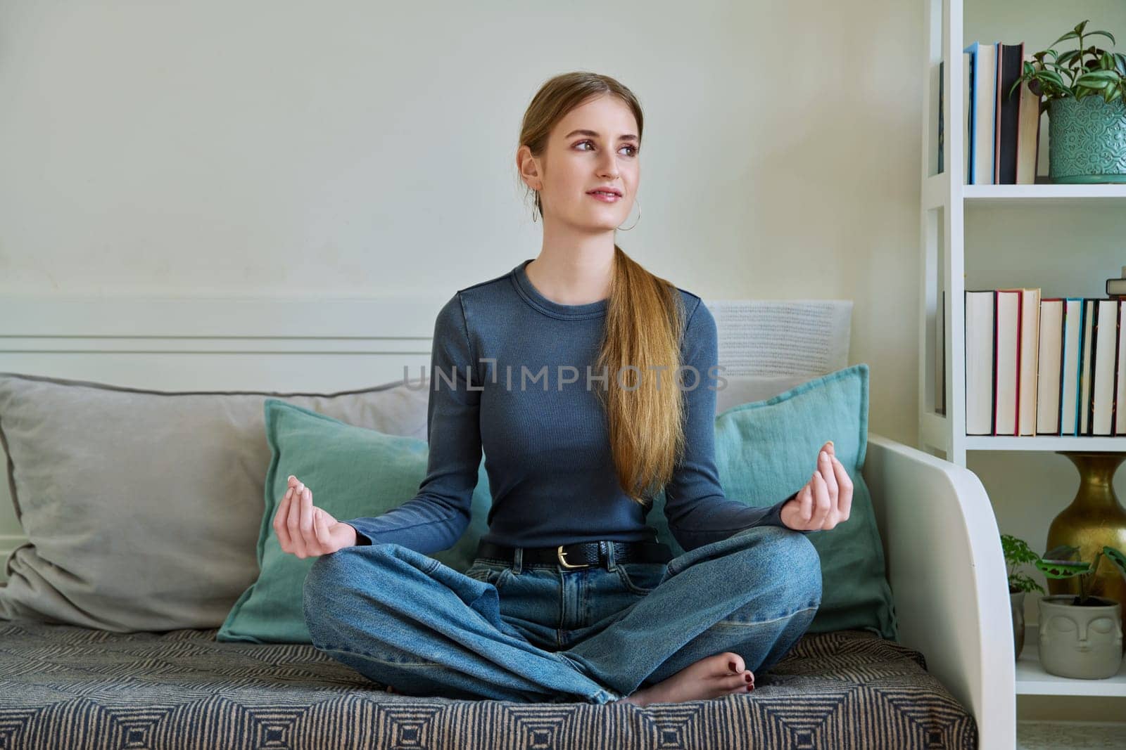 Young teenage calm relaxed female sitting in lotus position, meditating on couch at home. Health, mental and physical, lifestyle, youth concept