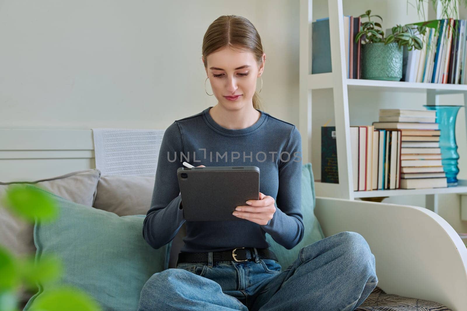 Young female teenager with digital tablet, stylus drawing illustrating sitting on couch at home. Technologies, leisure, creativity, education, freelance work, lifestyle, youth concept