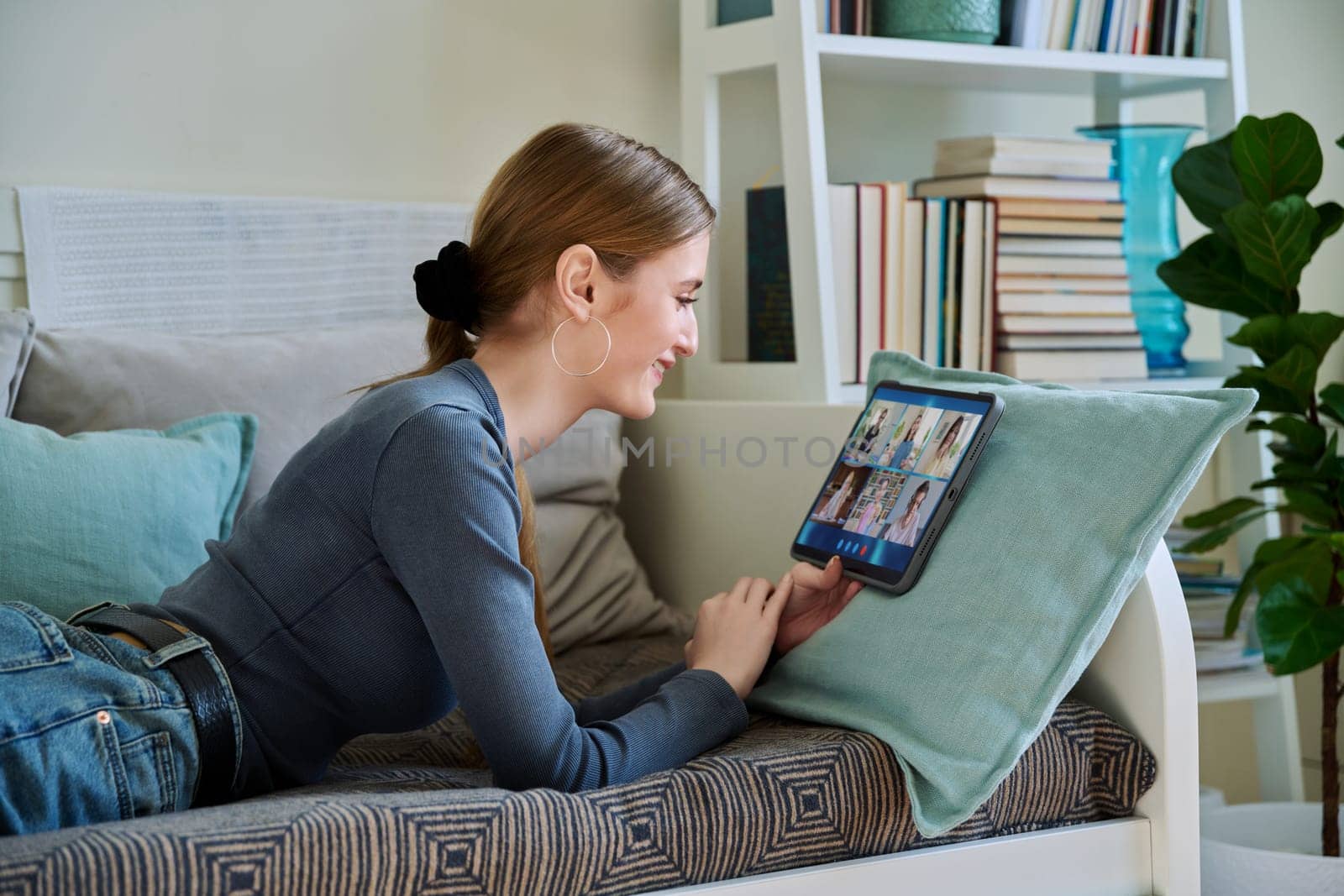 Female teenage student having group video conference, using digital tablet, lying on couch at home. by VH-studio