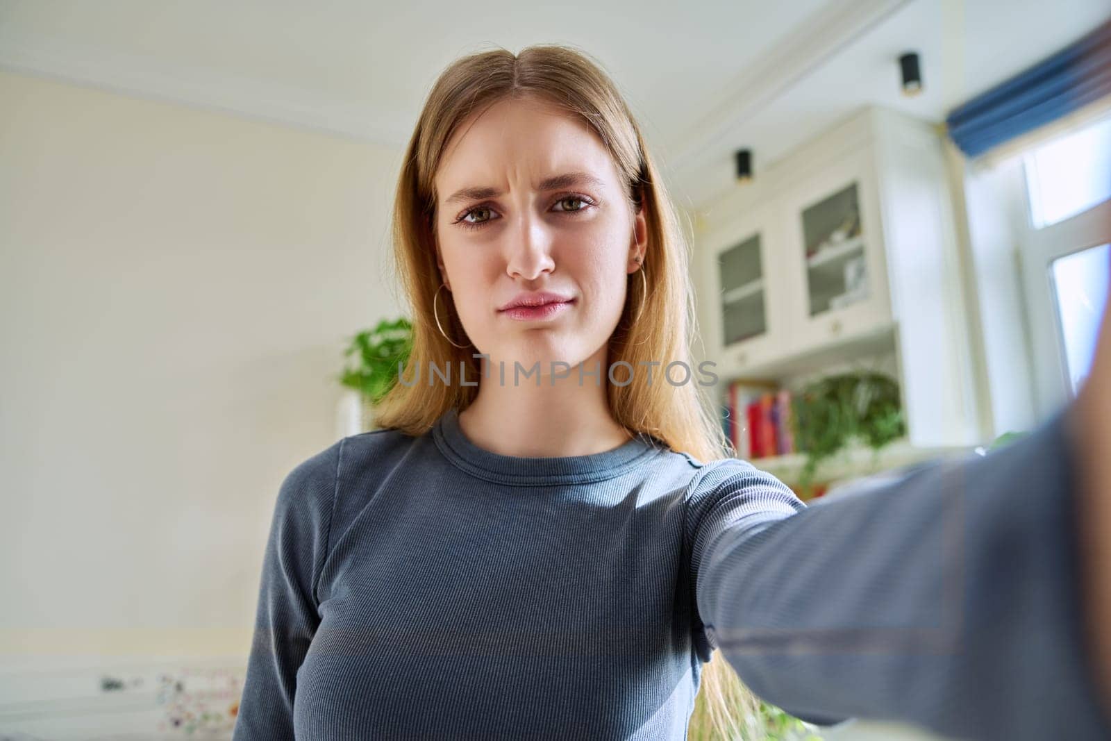 Close-up selfie portrait of teenage confused concentrated grimacing young female looking at web camera. Serious teenage girl 16, 17, 18 years old with blond hair, in home. Beauty, lifestyle, youth