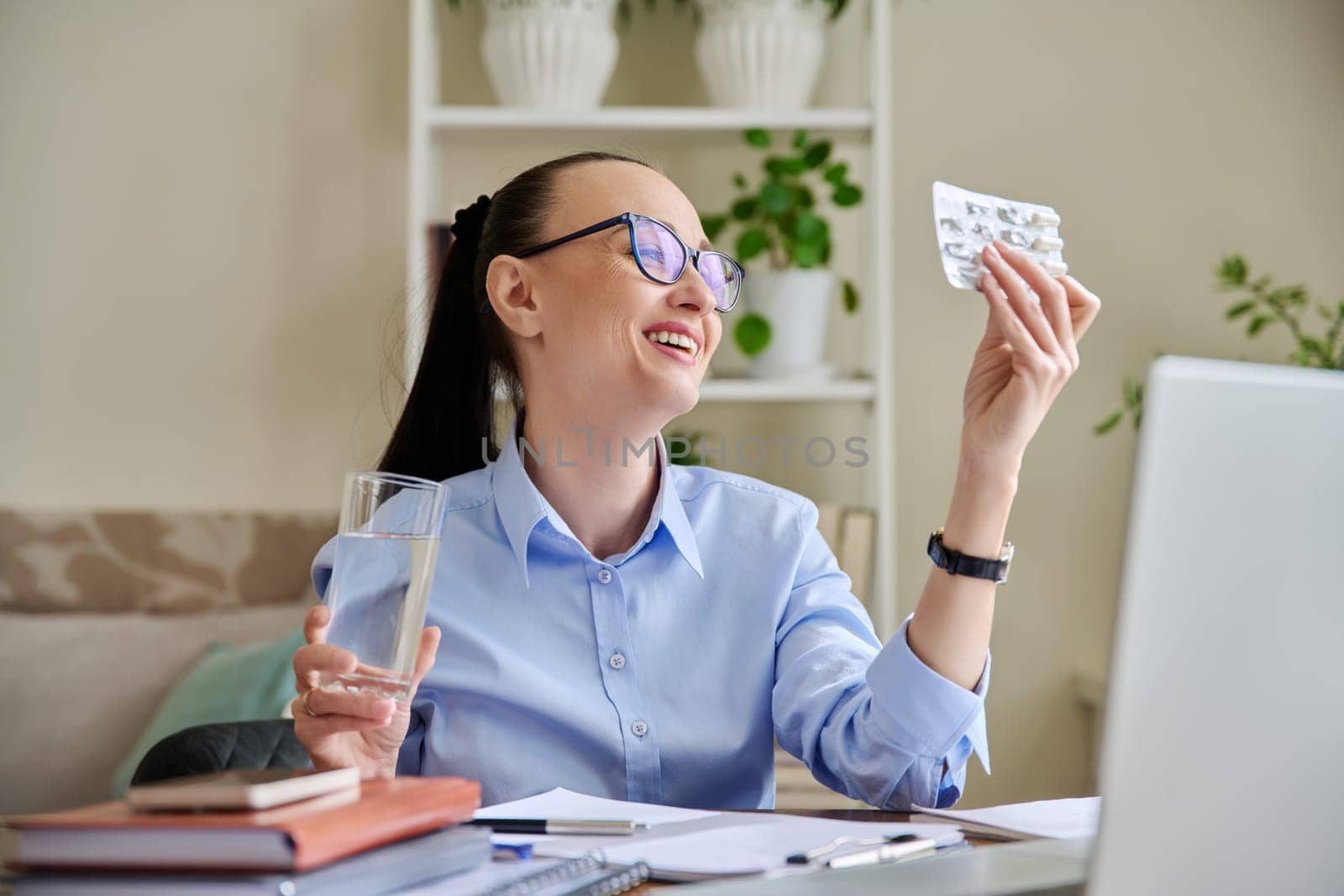 Happy woman at workplace holding blister with capsules glass of water. Female taking painkillers vitamins nutritional supplements antiviral drugs antioxidants antidepressants. Pharmacy health care