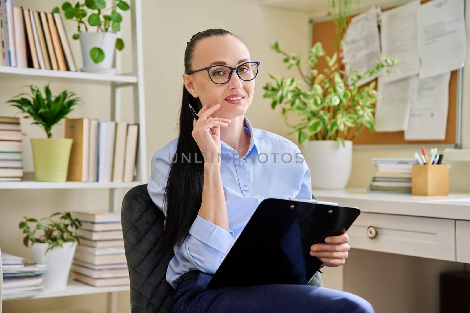 Portrait of smiling female psychotherapist with clipboard at workplace in office. Professional mental therapist counselor psychologist social worker looking at camera. Health care services, treatment