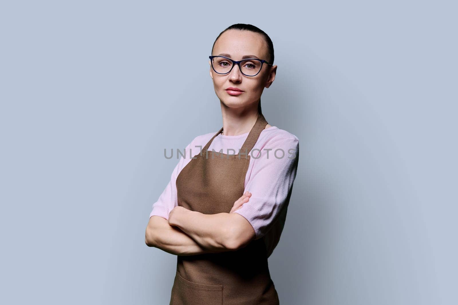 Portrait of confident serious 30s woman in apron on grey studio background. Successful female small business owner, service worker looking at camera with crossed arms. Staff, management, advertising