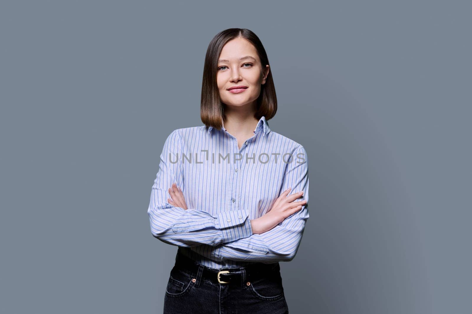 Young confident smiling business woman with crossed arms on grey studio background. Positive happy female in shirt, student worker owner entrepreneur looking at camera. Business work education people