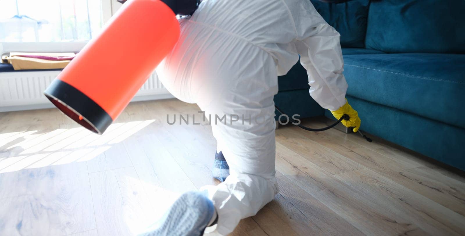 Worker in protective suit treating with disinfectant solution on floor under sofa in apartment by kuprevich