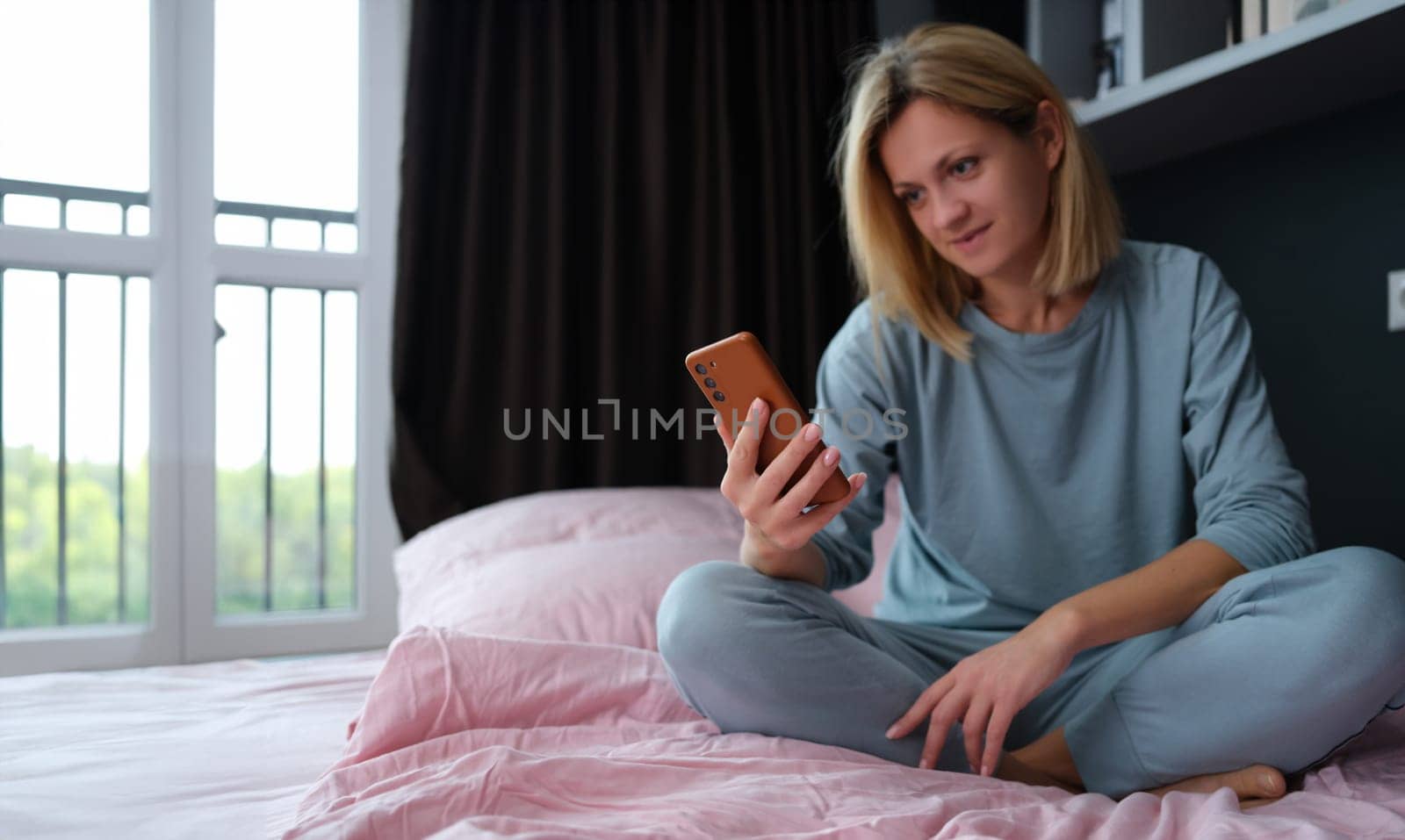 Young woman in pajamas sitting in bed with mobile phone in hand by kuprevich