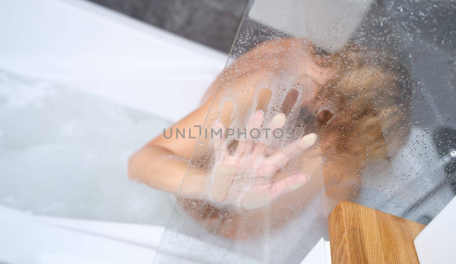 Naked woman sitting in bathtub and holding her hand on misted glass closeup by kuprevich