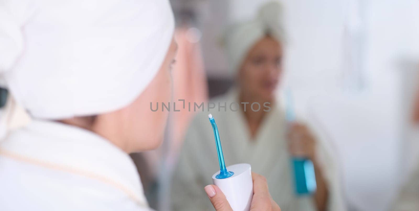 Young woman in gown and towel on head and brushing her teeth with irrigator in front of mirror in bathroom closeup by kuprevich