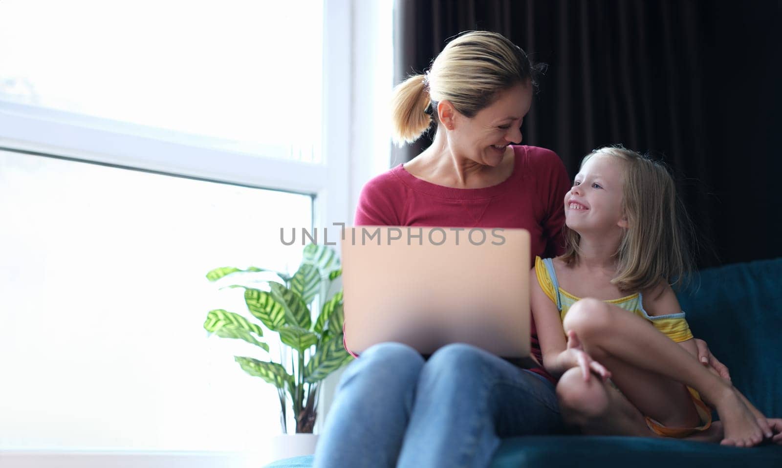 Little girl and mom are sitting on couch with laptop on laps and talking. Online education for children concept