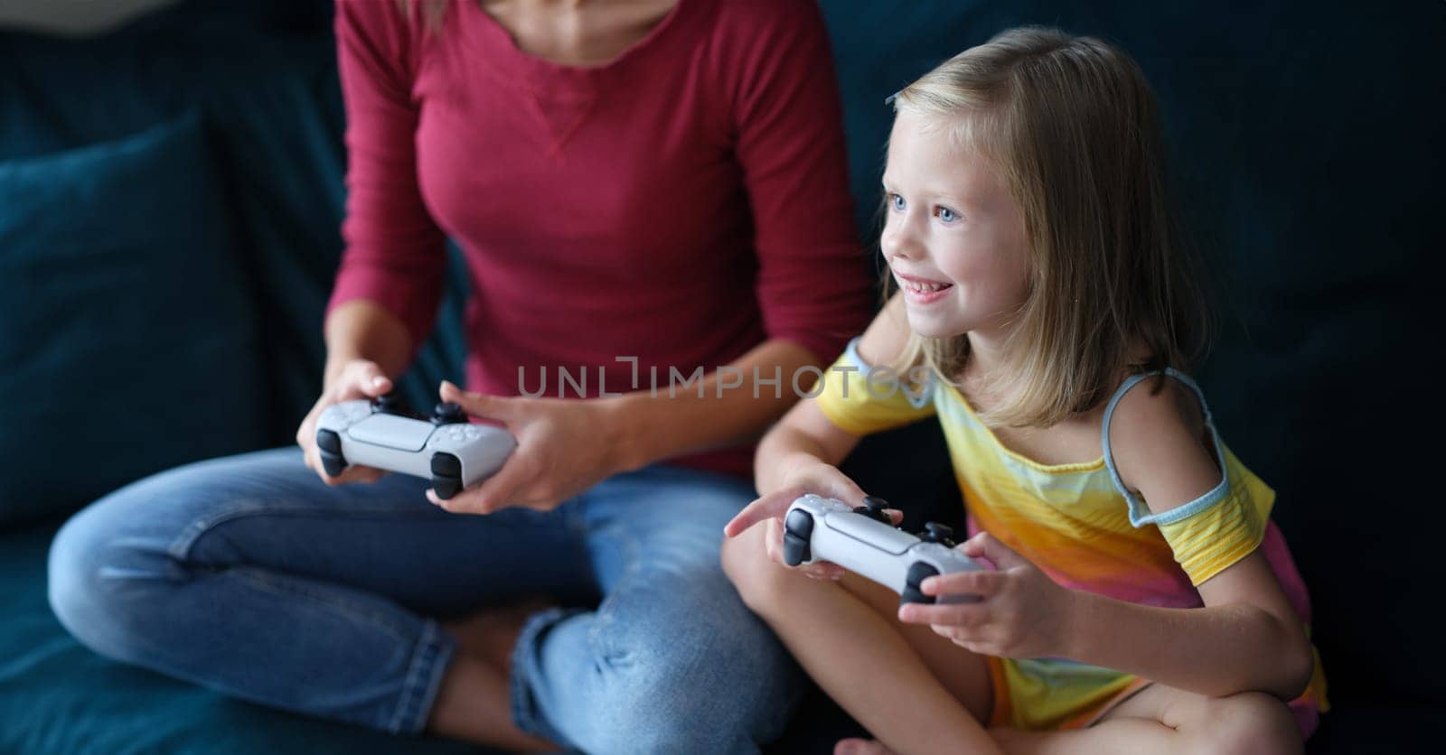 Little girl and mother sitting on couch and holding computer joysticks in their hands by kuprevich