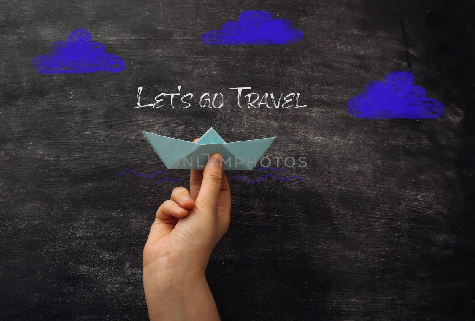 A hand holding a paper boat with the words Let's go travel written on a chalkboard