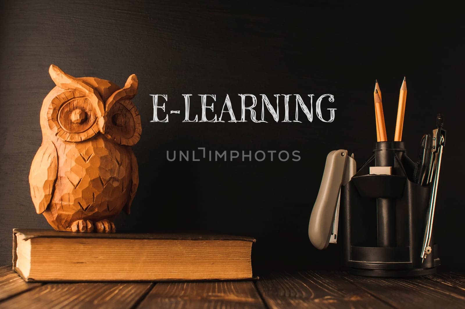 A wooden owl sits on top of a stack of books. The owl is surrounded by pencils and a pen