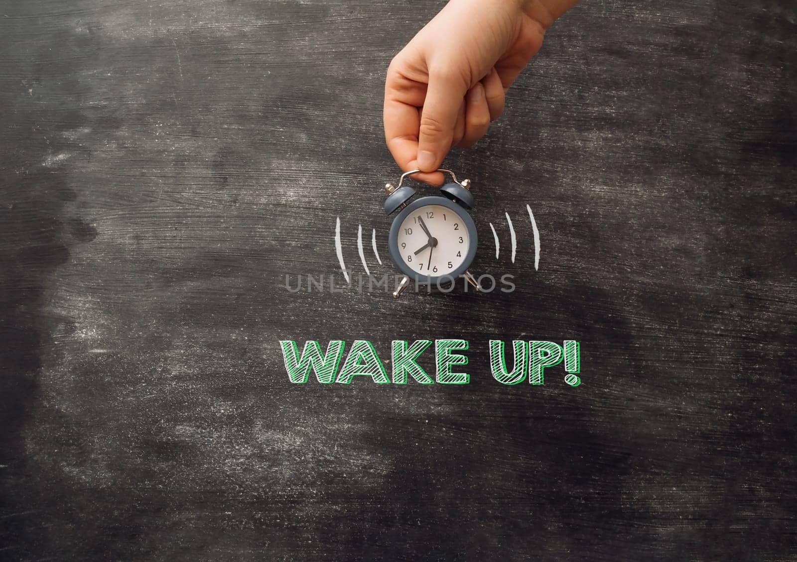 A hand holding an alarm clock with the words wake up written on the chalkboard. Concept of waking up early and the importance of starting the day on time