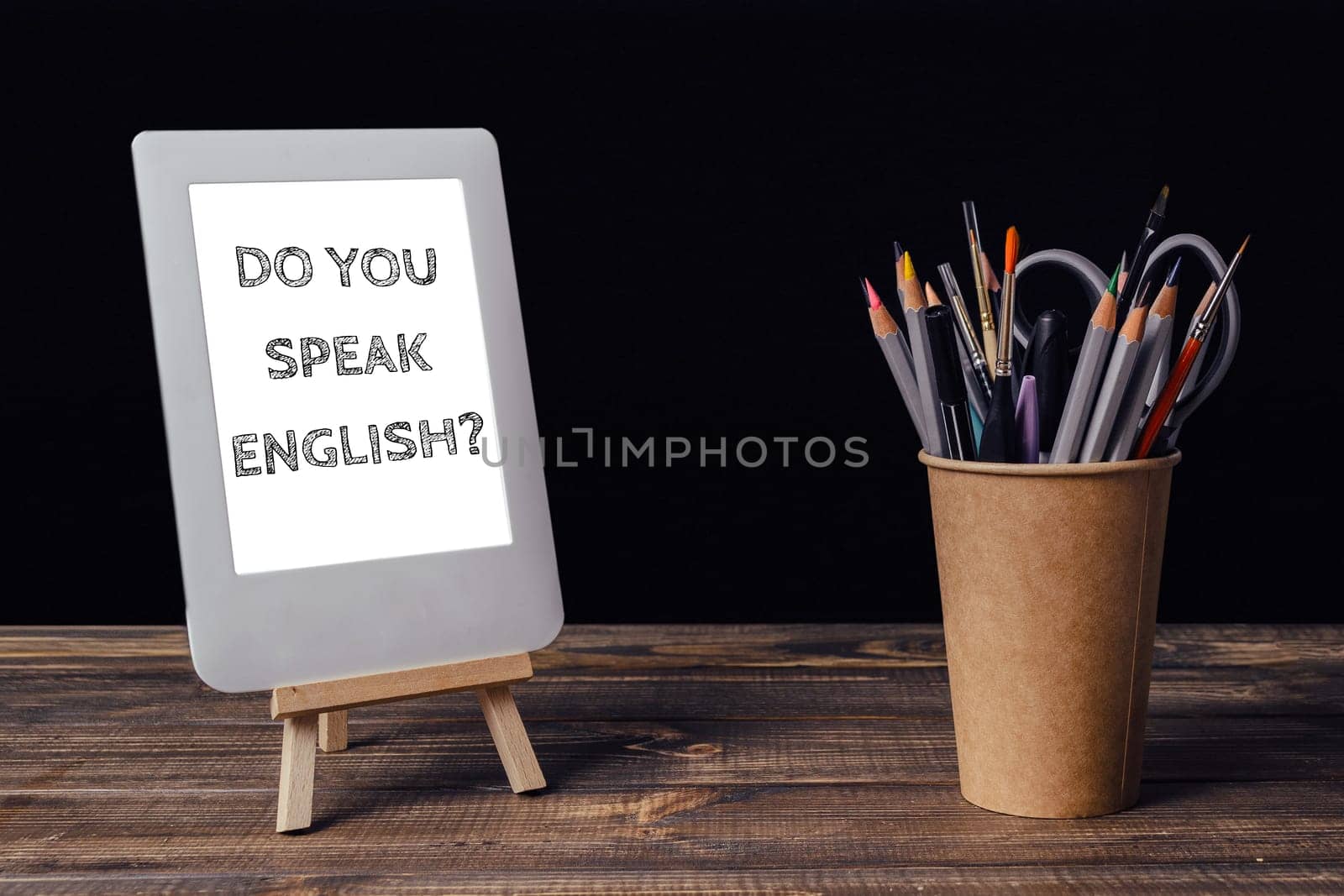 A white tablet with the words Do you speak English written on it. A pencil holder with a variety of colored pencils sits next to the tablet