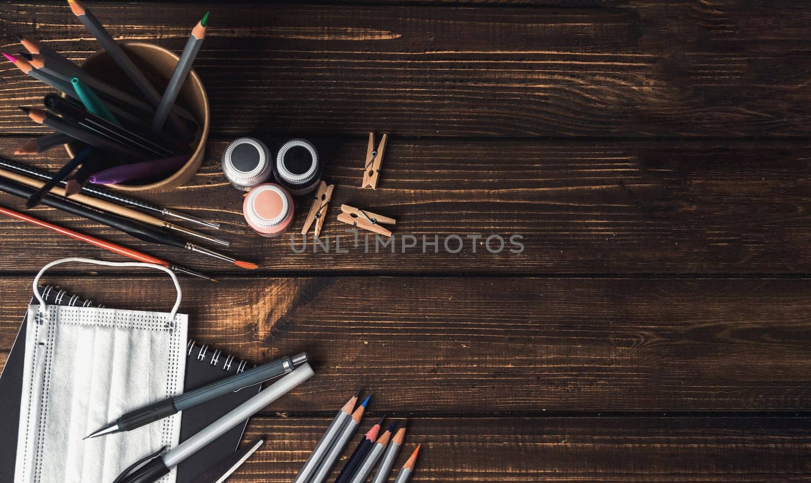A wooden table with a variety of art supplies and a face mask. The supplies include pencils, pens, and markers, while the face mask is placed on the table. The scene suggests a creative