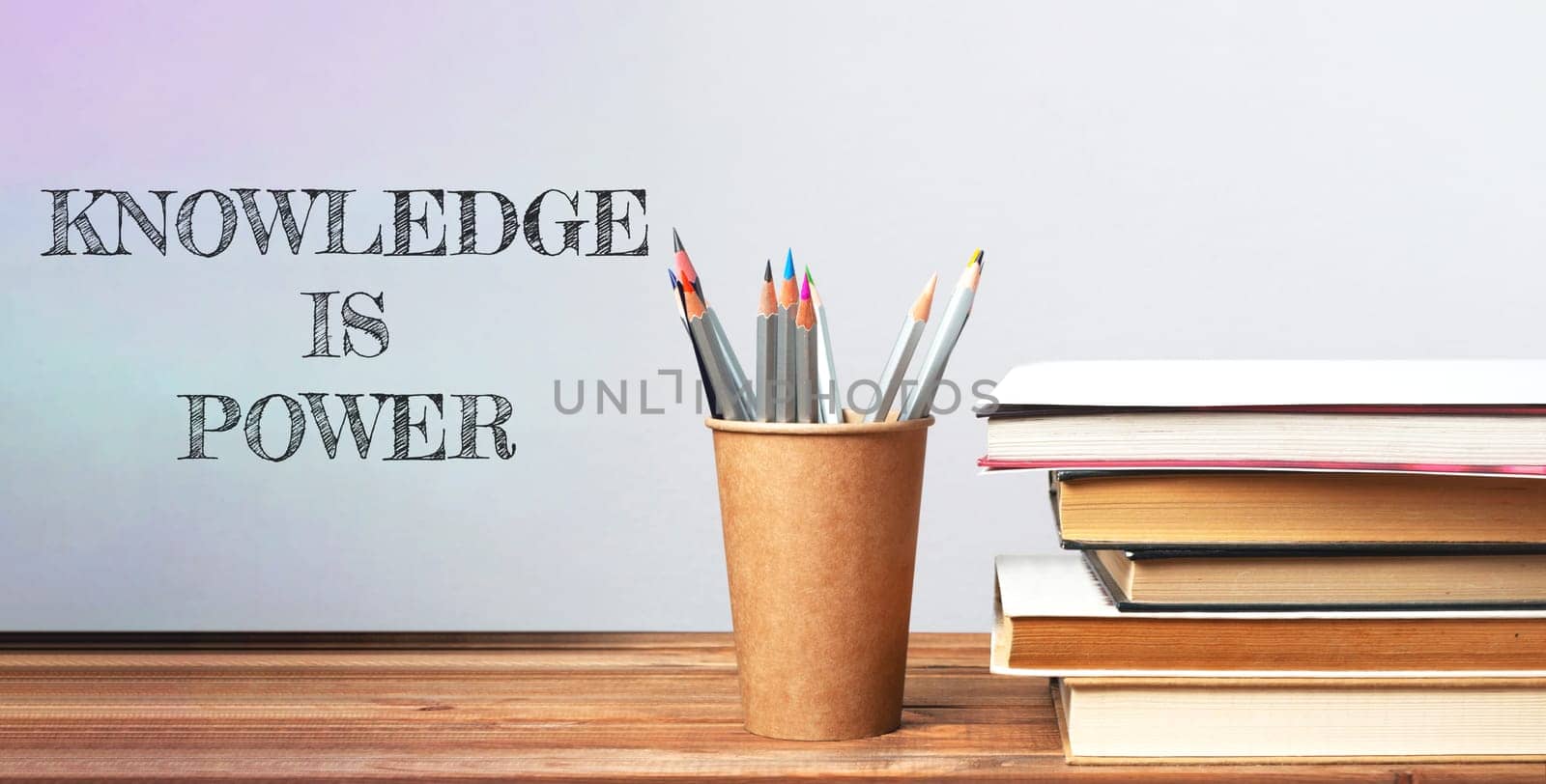 A wooden desk with a cup of pencils and a stack of books. The words knowledge is power are written on the wall behind the desk