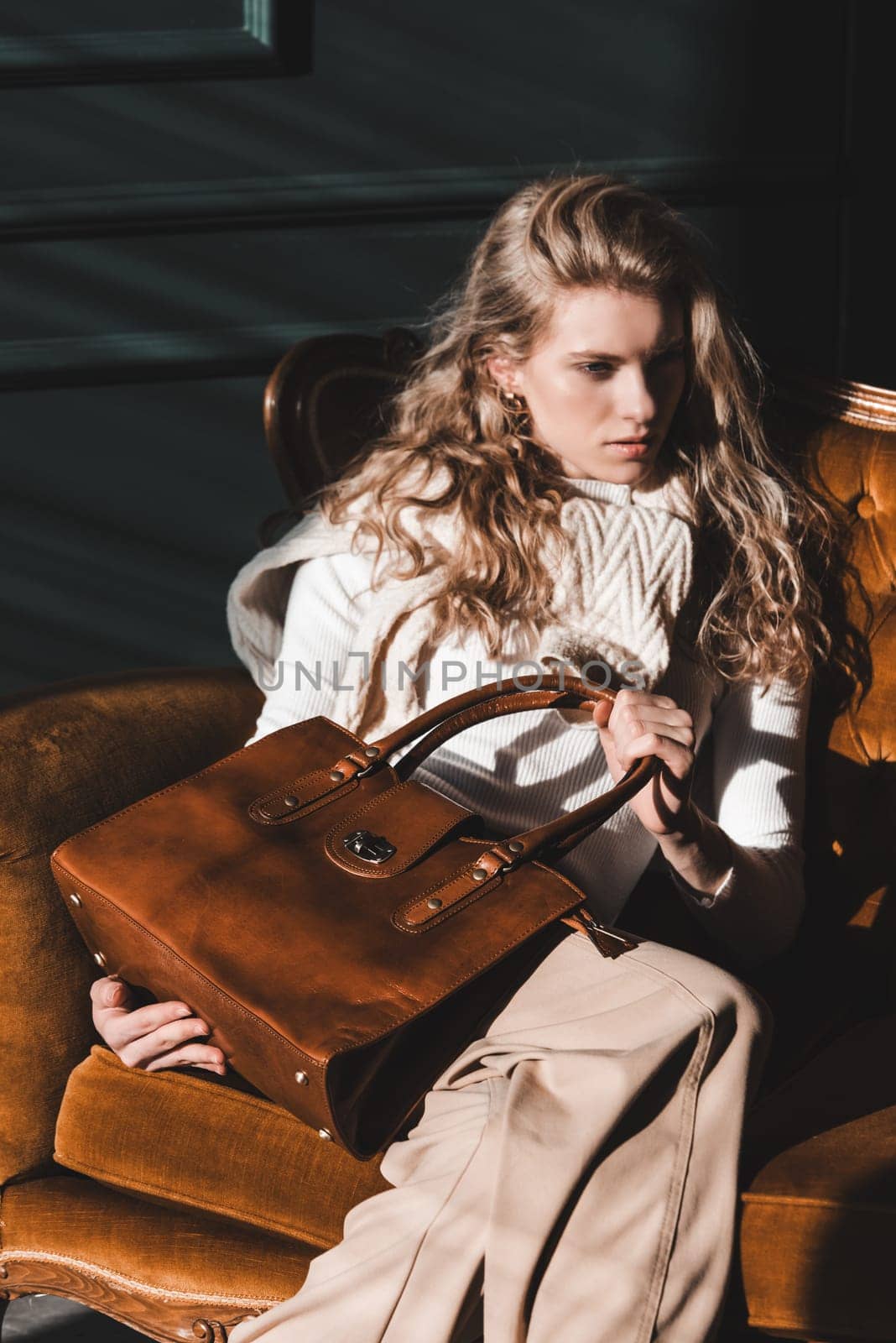 beautiful curly blond hair woman posing with a small shopper brown bag in a vintage chair. Model wearing stylish white sweater and classic trousers