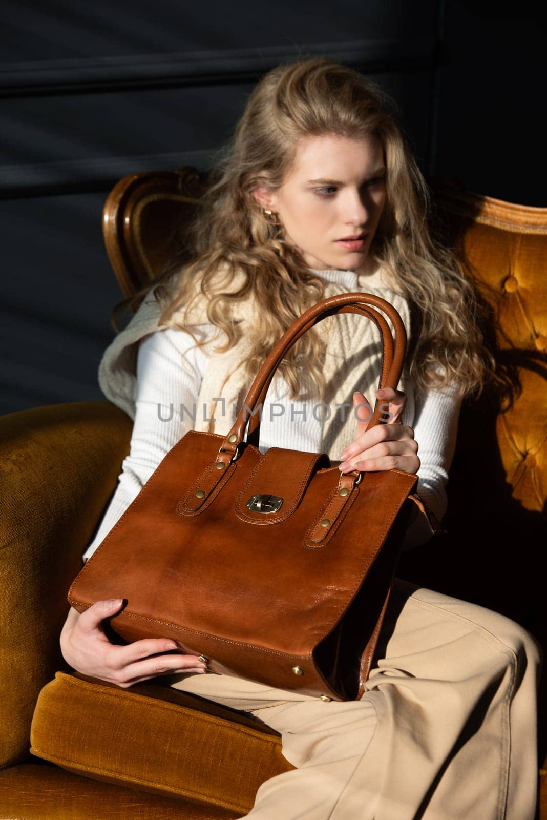 beautiful curly blond hair woman posing with a small shopper brown bag in a vintage chair. Model wearing stylish white sweater and classic trousers