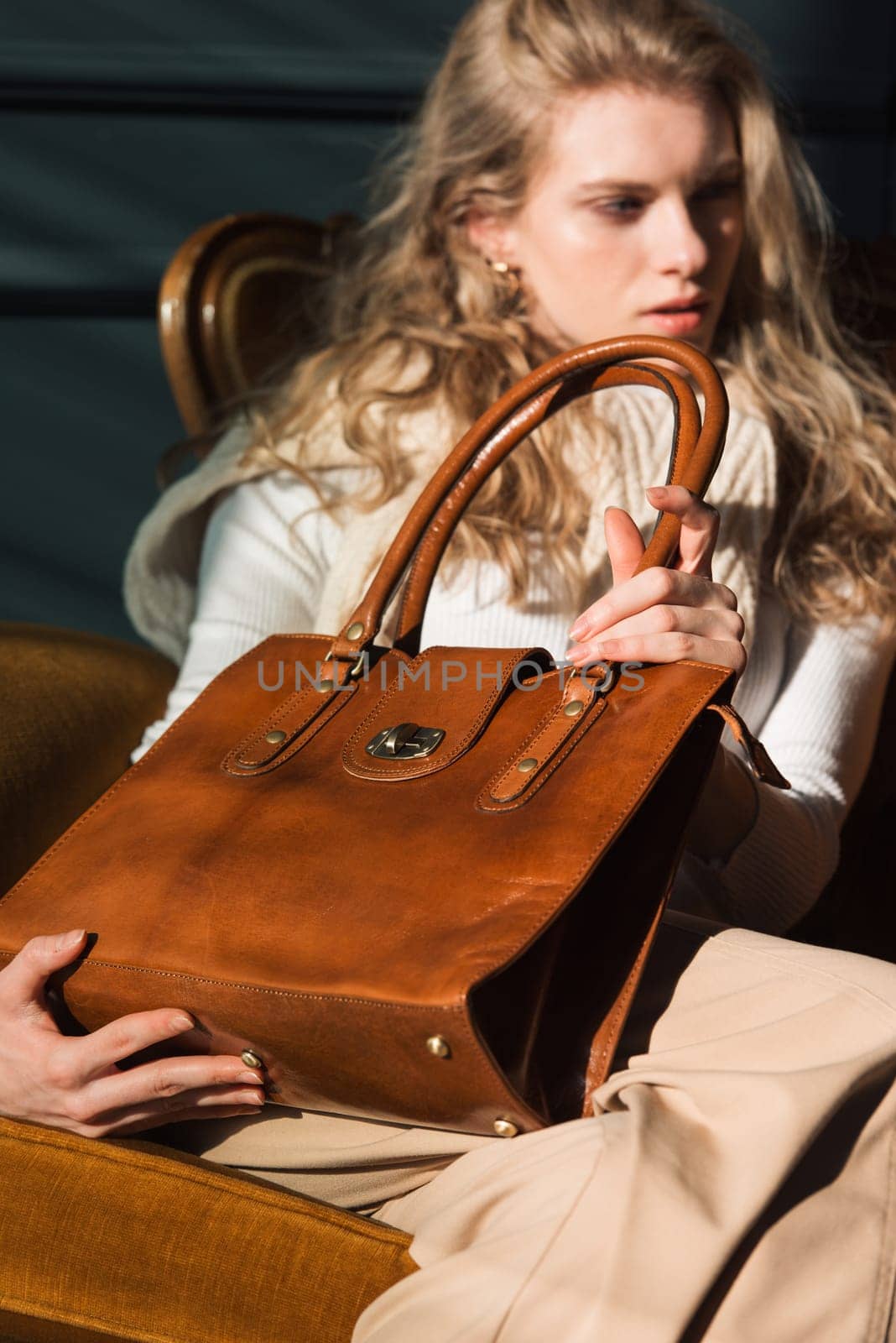 beautiful curly blond hair woman posing with a small shopper brown bag in a vintage chair. Model wearing stylish white sweater and classic trousers