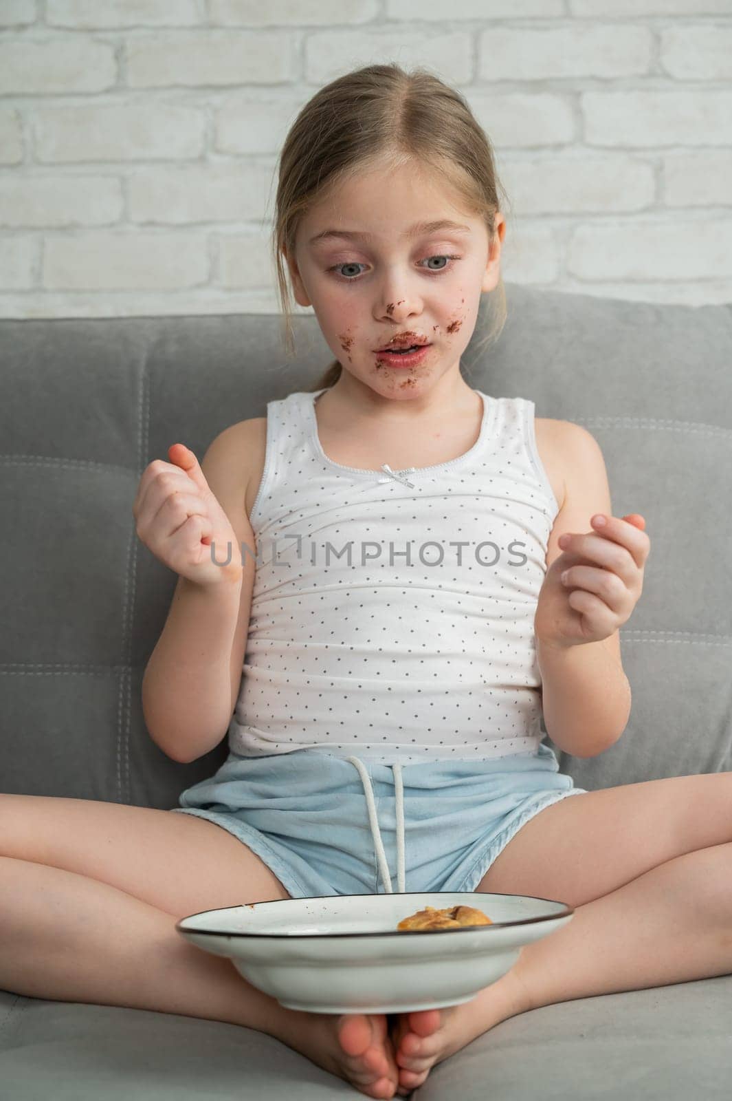 A cute little girl covered in chocolate eats cookies while sitting on the sofa. Vertical photo. by mrwed54