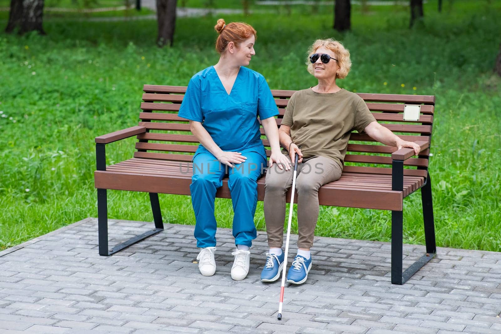 A nurse and an elderly blind woman are sitting on a bench in the park. by mrwed54