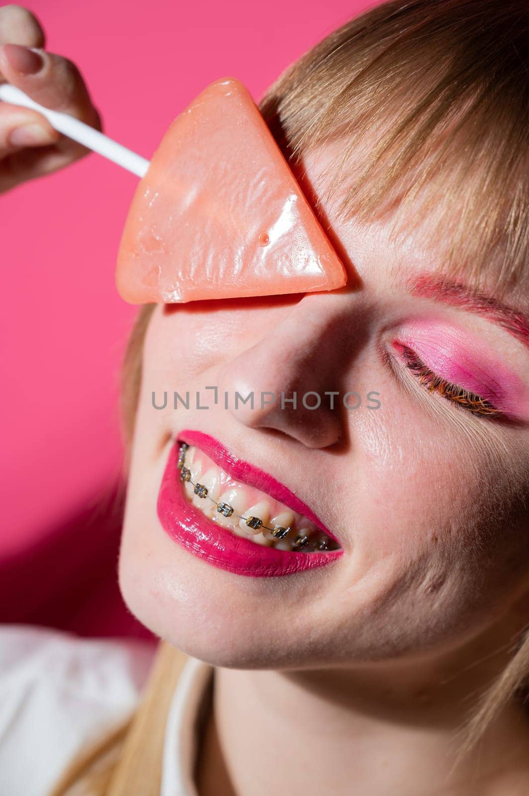 Portrait of a young woman with braces and bright makeup eating a lollipop on a pink background. Vertical photo. by mrwed54