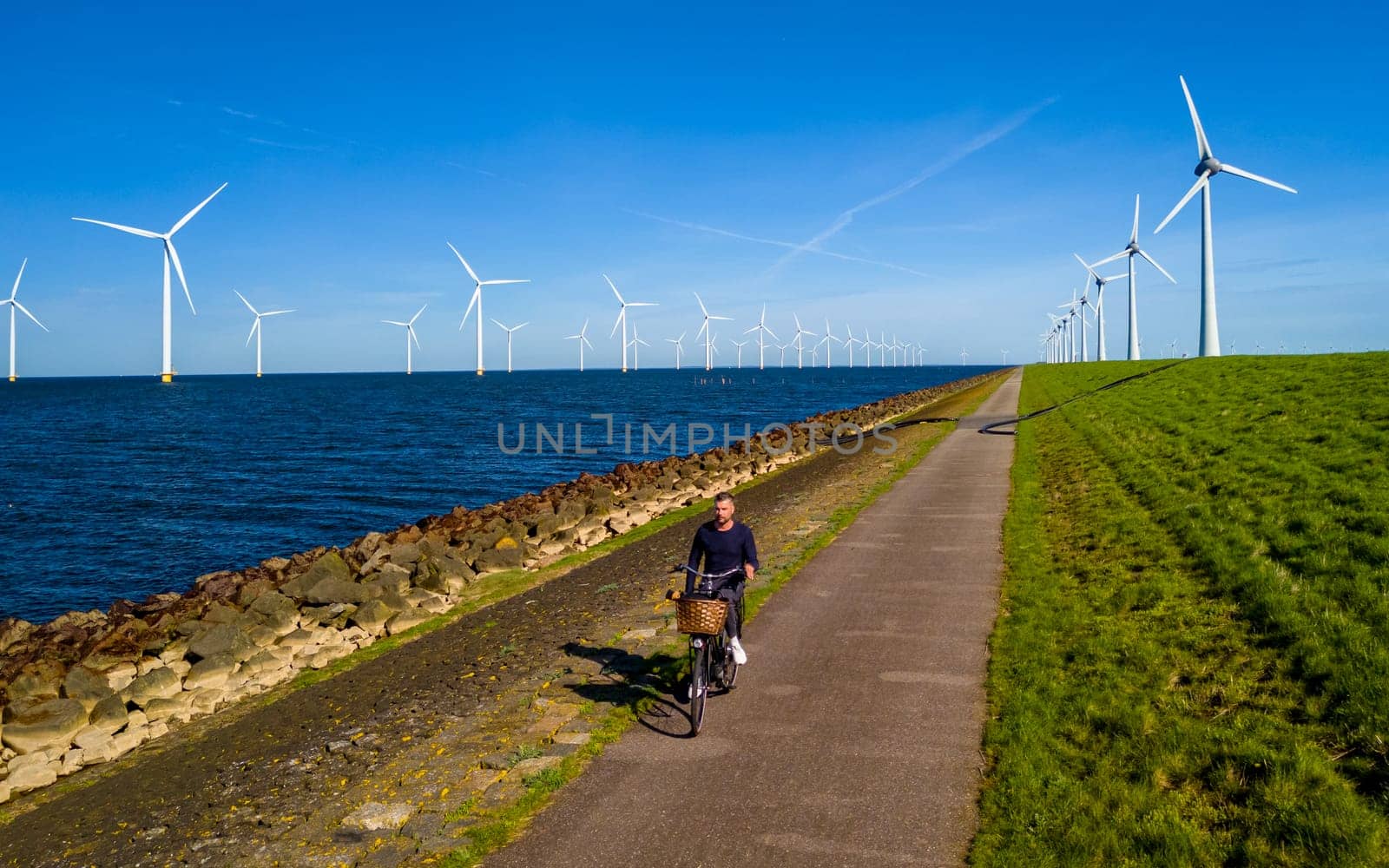 A man is peacefully cycling down a serene path next to a body of water in Flevoland, Netherlands. The scene is surrounded by windmill turbines, creating a picturesque and idyllic setting for a leisurely bike ride.