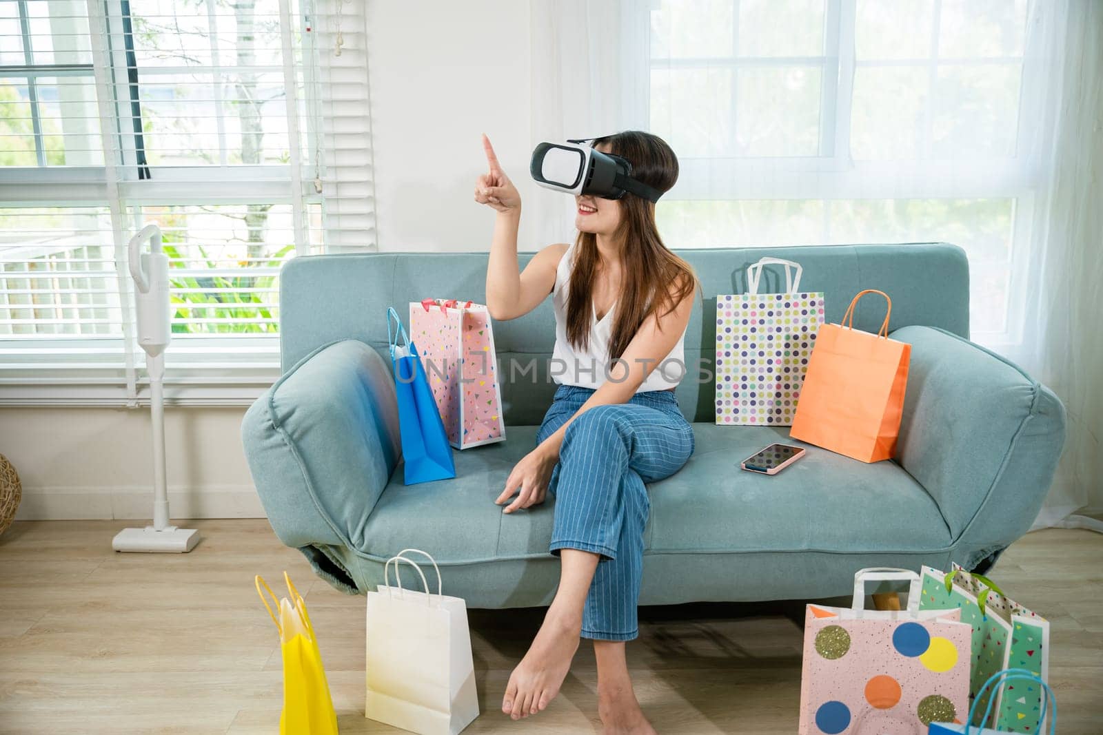 Happy young woman customer wearing 3d VR glasses virtual reality headset with shopping bags around she touching and pointing on air to online shopping in living room at home, digital cyberspace