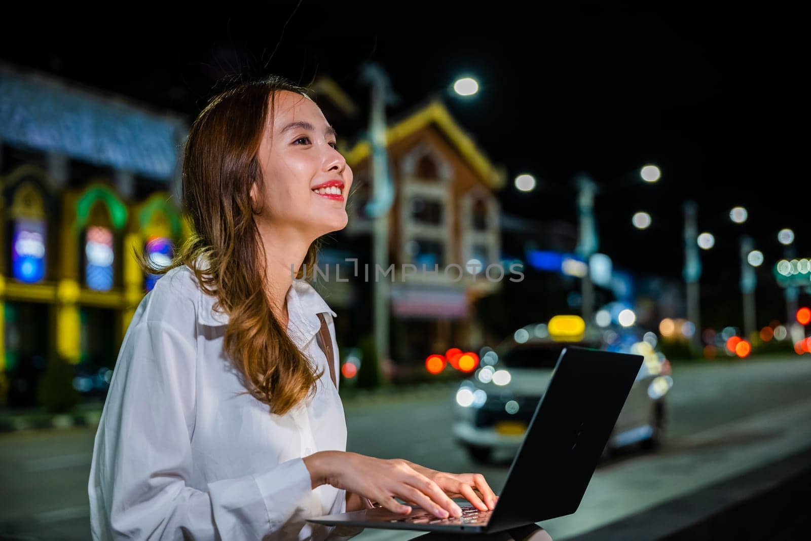 On a bustling city street at night, a woman is hard at work on her laptop computer, utilizing the convenience and mobility of her device to stay connected in the digital age.