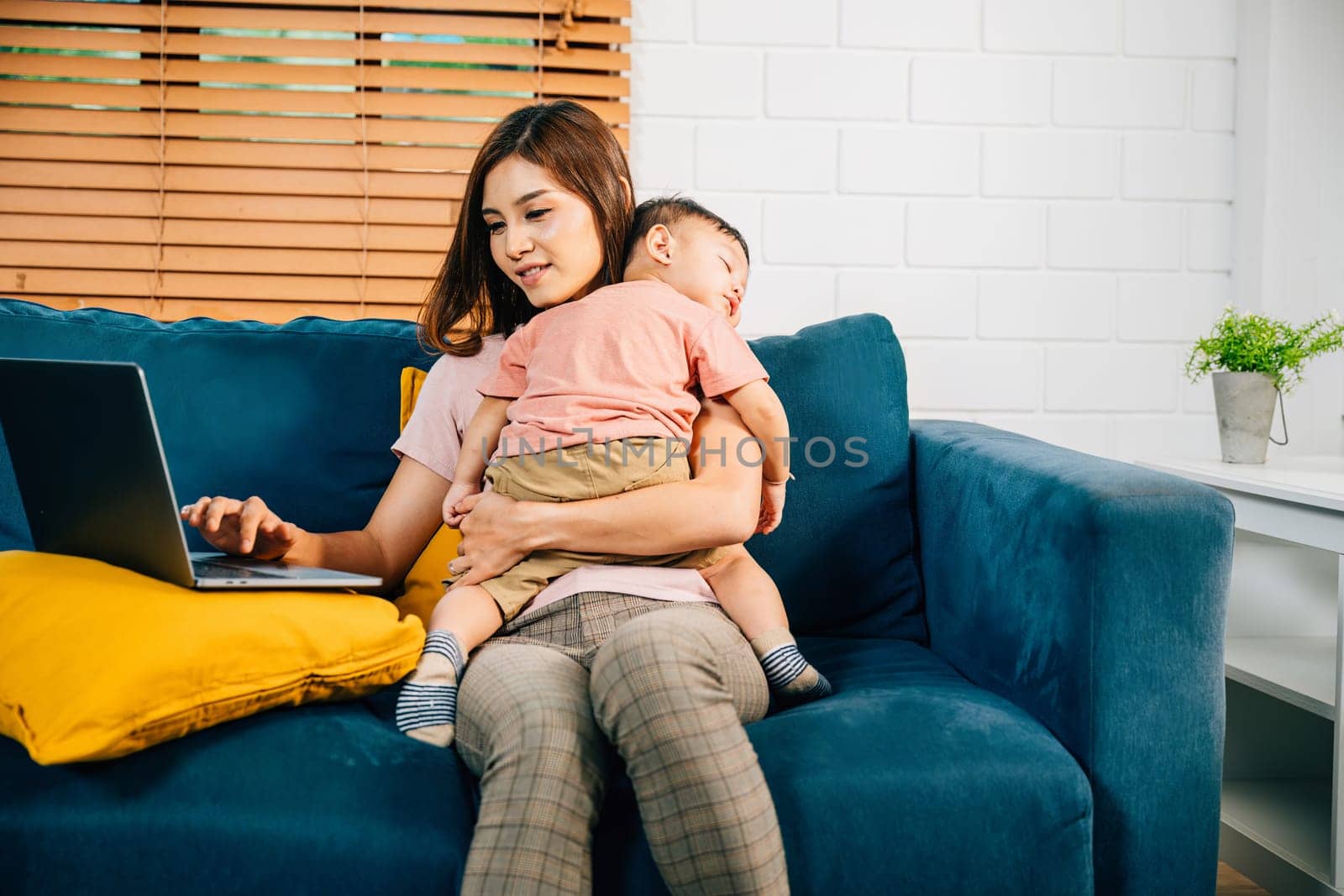 Balancing business and family a mother types on her laptop as her baby daughter naps on the sofa. The affectionate care and responsibility are beautifully captured in this portrait.