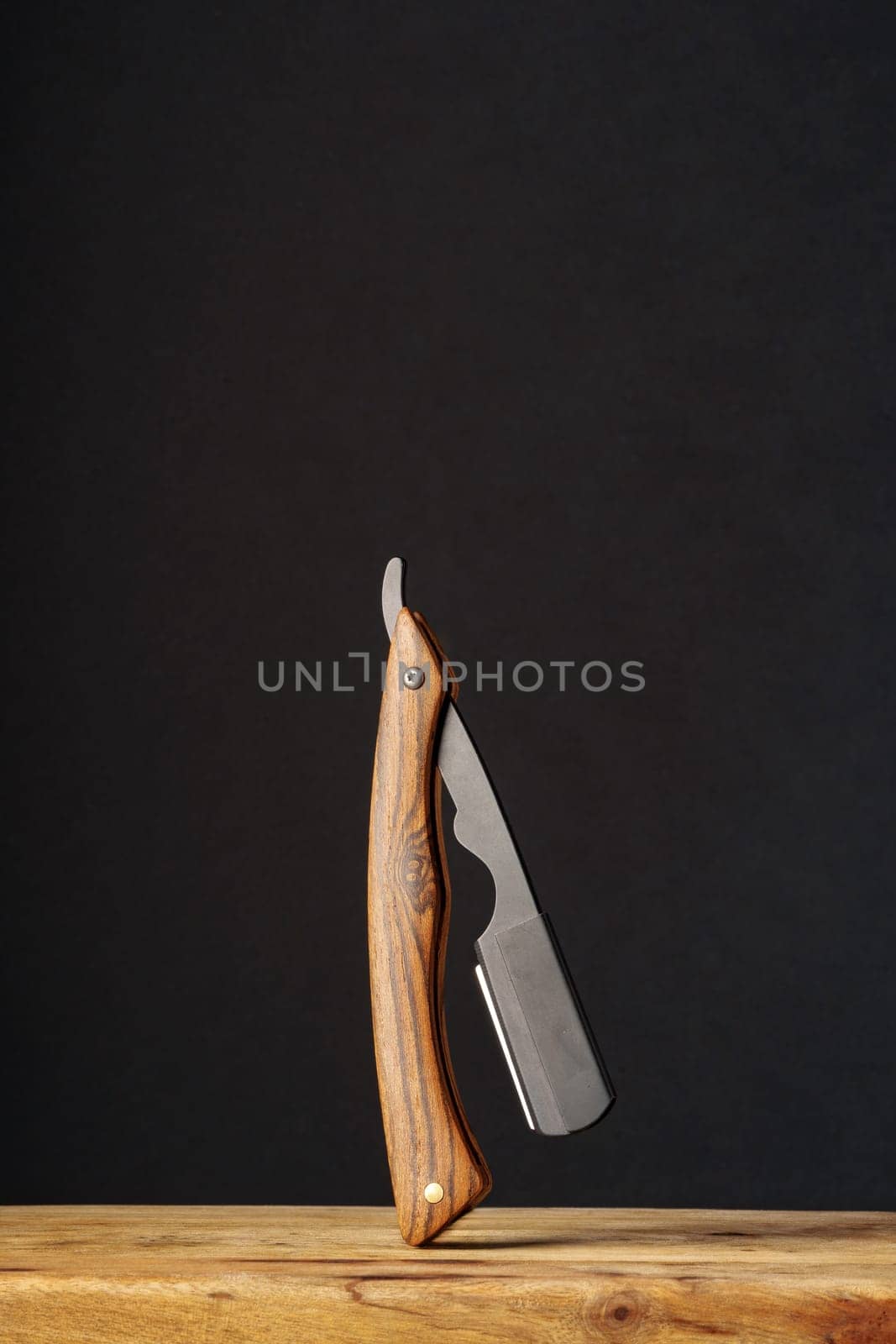 A sleek folding knife with a wooden handle is showcased in an upright position on a rustic wooden platform, presenting a contrast against the stark, dark backdrop.