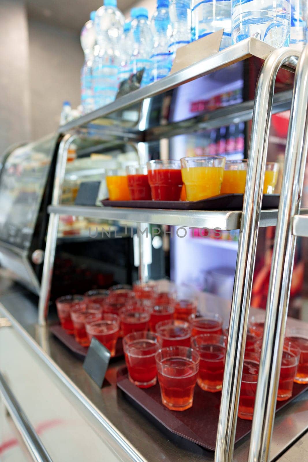 A selection of fruit juice cups, with orange and red hues, are neatly arranged on a metal serving cart, ready for patrons to enjoy in a bustling cafeteria setting. The focus on the front cups, with a blurred background, highlights the refreshment options available.