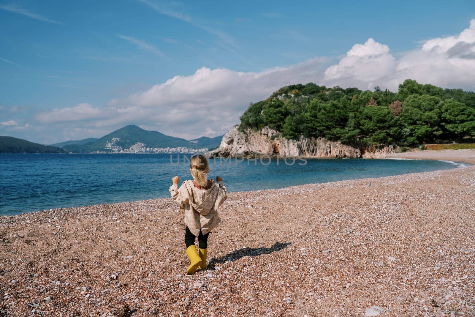 Little girl walks along the beach to the sea looking at the mountains. Back view. High quality photo
