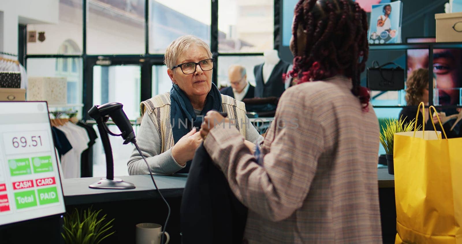 African american employee scanning clothes tags at checkout, preparing bag with products for elderly woman at department store. Retail worker using scanner at cash register, consumerism. Camera B.