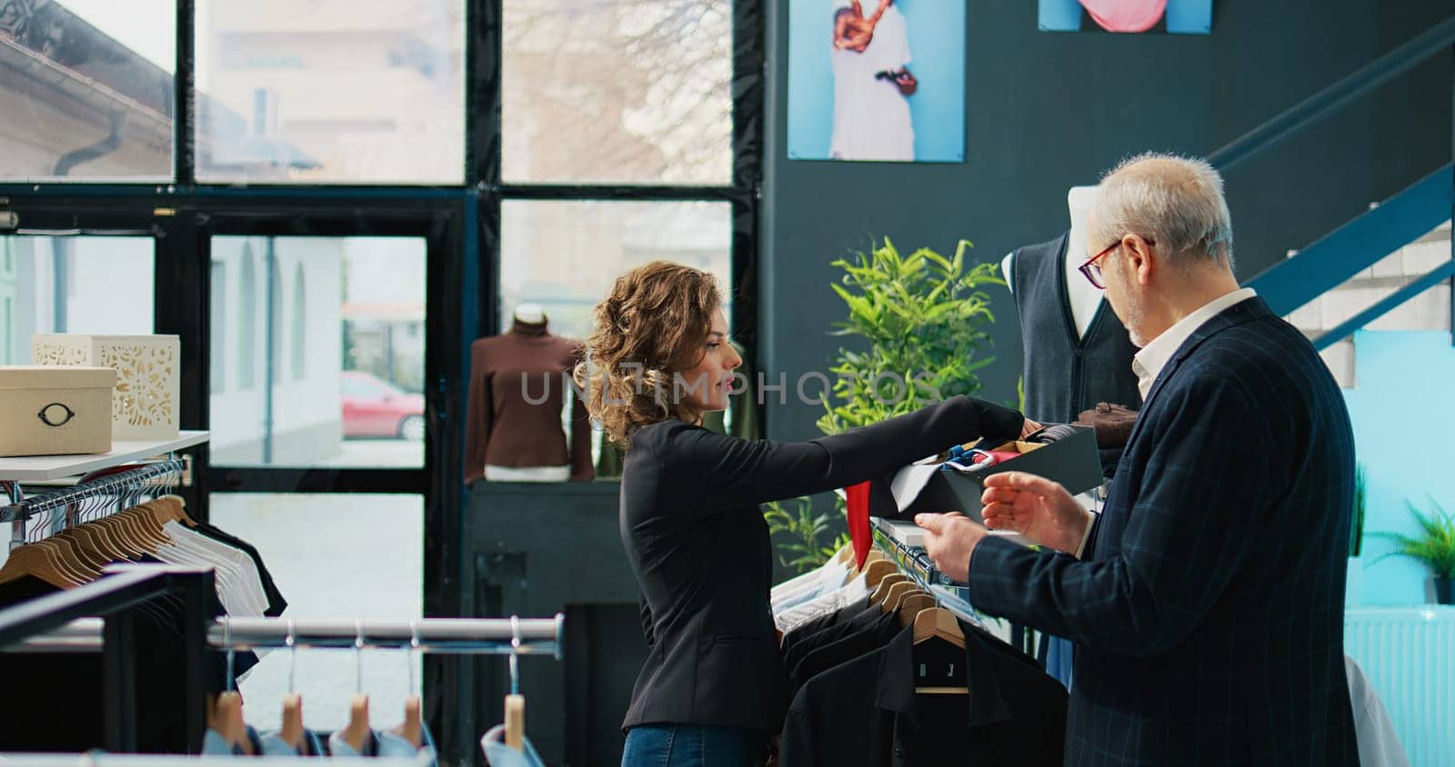 Woman employee presenting formal accessories to regular client, assisting him in choosing the right tie color to match his suit jacket. Store clerk helping customer, commercial activity. Camera B.