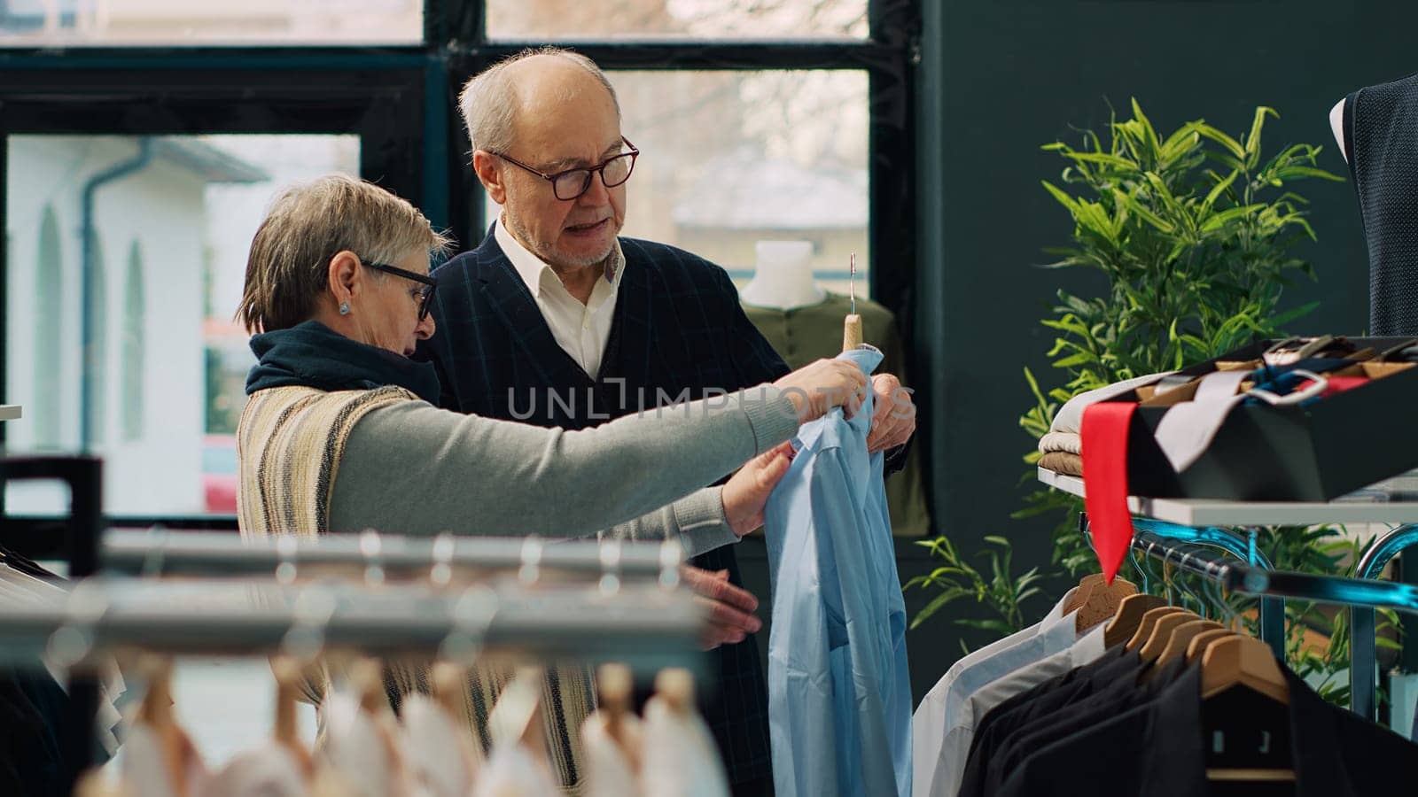 Senior couple shopping for formal outerwear at fashion boutique, looking for trendy elegant clothing items to create new outfits for elderly man. People searching for stylish merchandise. Camera A.