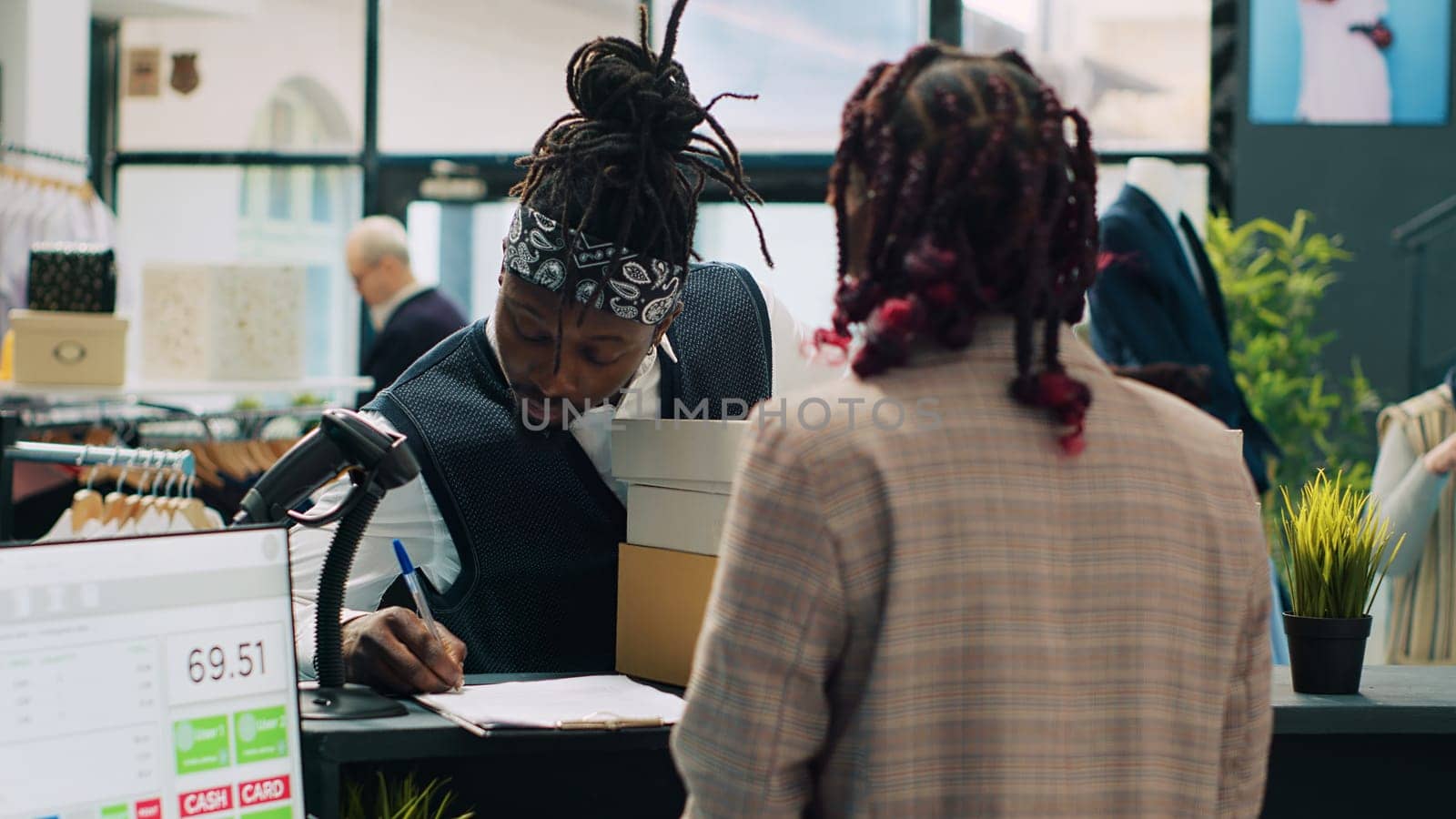 African american deliveryman receiving requested clothes packed in boxes, arriving at fashion showroom to pick up order and sign papers. Woman employee preparing items at cash register. Camera A.