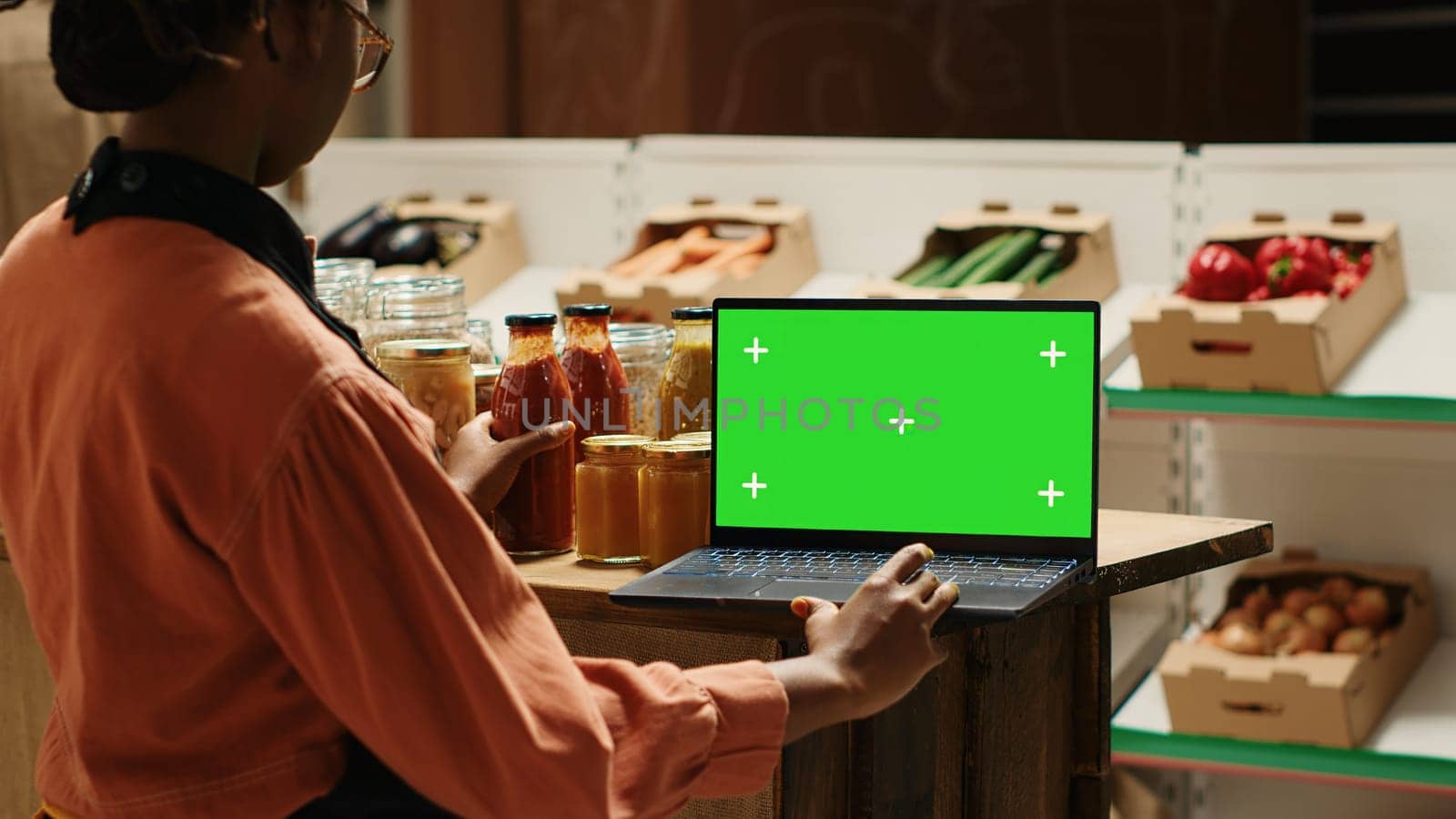 Young vendor looks at greenscreen on laptop while preparing merchandise for customers, using isolated mockup layout on screen. African american seller checking blank chromakey display. Camera 1.